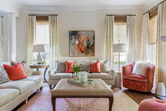 Feminine Living Room with neutral shades and drapery, neutral furniture with coral accents, leather ottoman, distressed pink rug, and original artwork.