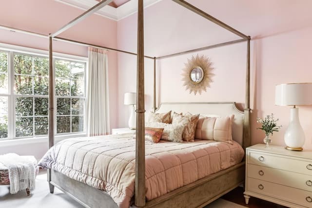 Feminine Bedroom with pink walls, four poster canopy bed, cream nightstands and neutral drapery.