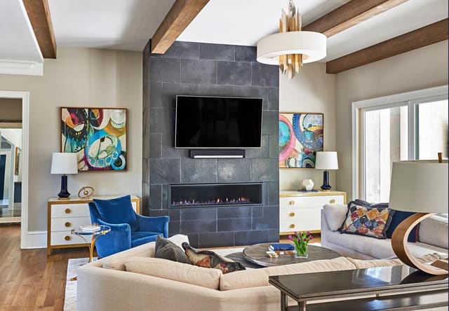 Family Room in Wake Forest home with exposed beams, horizontal fireplace, colorful artwork, blue velvet chair, twin ivory and gold chests, and relaxed sectional.