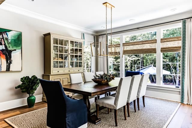 N. Raleigh Breakfast room with sisal rug, hanging pendant, dark wood table, white upholstered dining chairs, navy skirted hostess chairs, woven wood shades, and custom window treatments