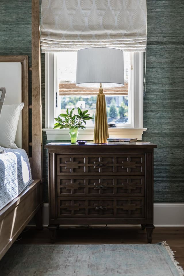 Master Bedroom close up showing chest with brass lamp, area rug, green raffia wallpaper, and neutral roman shade.
