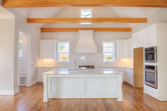Kitchen in North Raleigh home with exposed wood beams, white cabinets, white range hood, and large island with white quartz countertop.