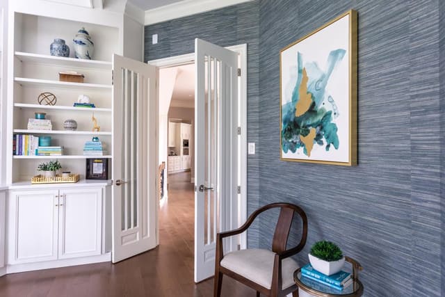 Blue grasscloth wallpapered office with framed abstract art and a decorated bookshelf.
